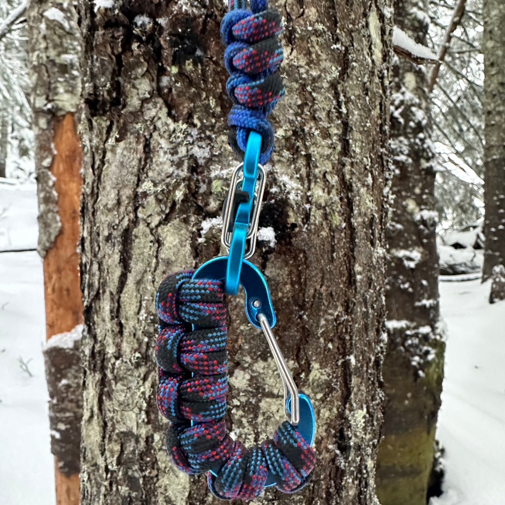 Multicolored paracord bracelet with carabiner attached to a tree in a snowy forest.